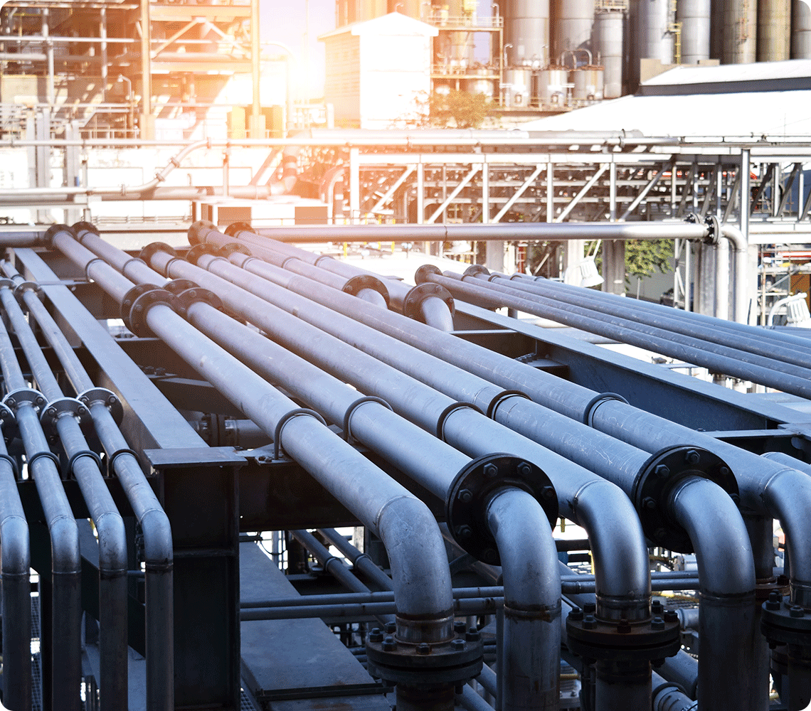 A network of industrial metal pipelines is shown outdoors at a facility, with buildings and additional industrial structures in the background. Sunlight is casting a glow on the upper left corner.